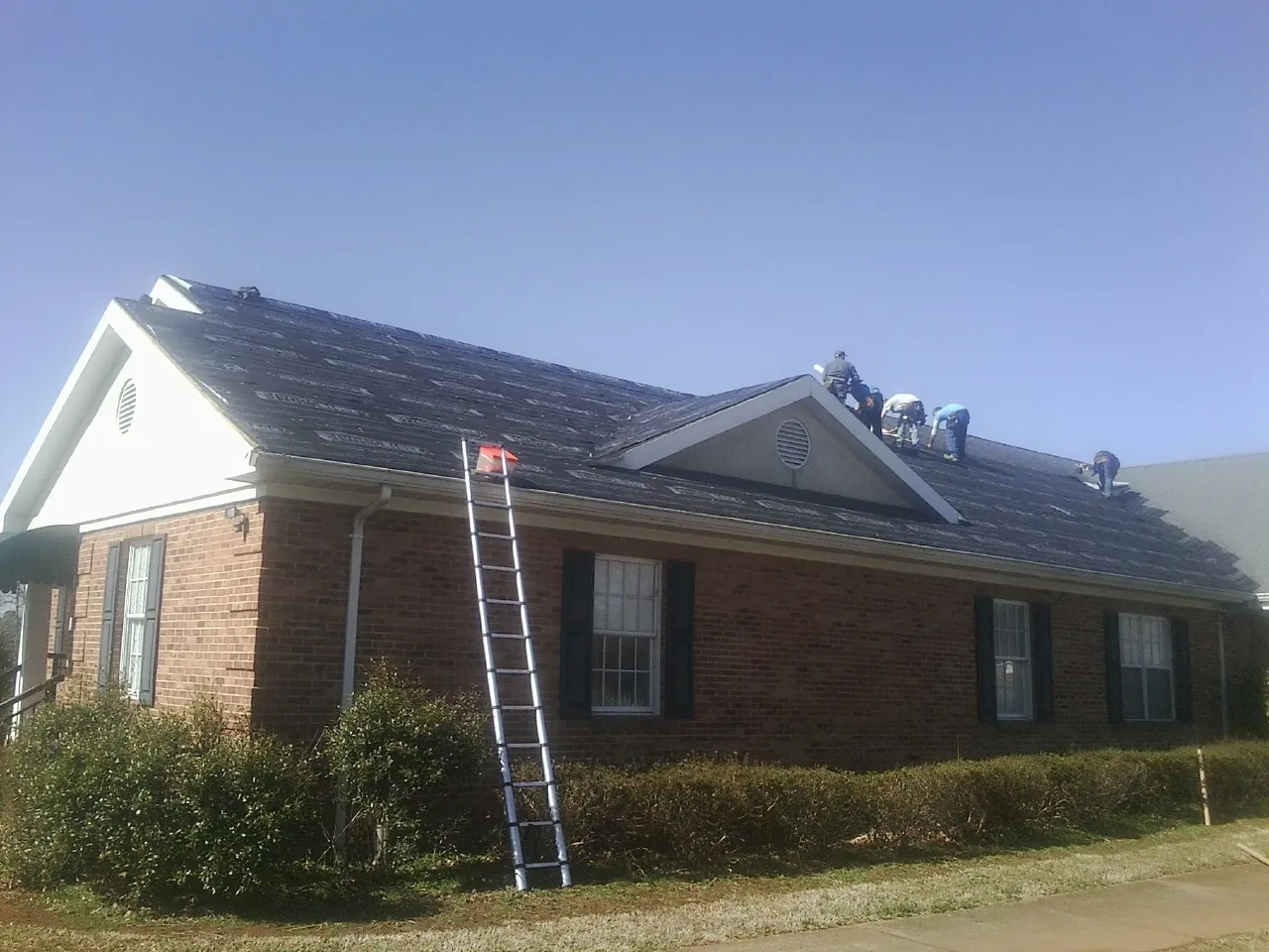 Crew tearing shingles off a brick ranch roof with a ladder set — Metal Roof Repair work by our crew in Camas Valley