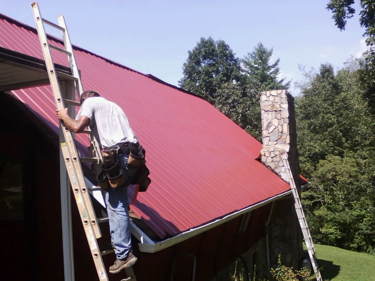 Roofer on a ladder beside a red metal roof and stone chimney — Soffit & Fascia Repair by Roofing Brothers in Camas Valley
